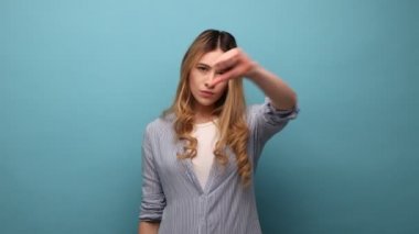 Mutsuz, tatminsiz genç bir kadının portresi baş parmaklarını aşağı gösteren, olumsuz duygular, kötü derecelendirme, çizgili gömlek giyen. Mavi arka planda kapalı stüdyo çekimi.