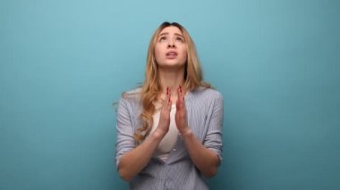 Dalgalı saçlı bir kadın dua ederken el ele tutuşuyor ve yalvarıyor, umut dolu gözlerle bakıyor, özür diliyor, çizgili gömlek giyiyor. Mavi arka planda kapalı stüdyo çekimi.