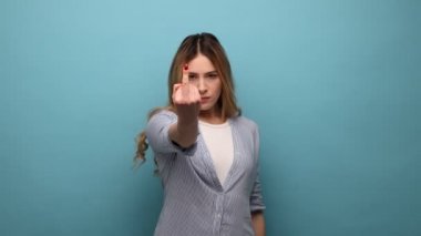 Sarışın kadının iletişimi reddetmesinin portresi, saygısızlığı ve nefreti ifade etmek için orta parmağını göstermesi, kaba bir jest, çizgili gömlek giymesi. Mavi arka planda kapalı stüdyo çekimi.