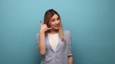 Beni ararsın. Dalgalı saçlı, el hareketleriyle ayakta duran kameraya gülümseyen, telefonla temas teklif eden, çizgili gömlek giyen bir kadın. Mavi arka planda kapalı stüdyo çekimi.