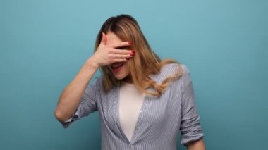 Güzel sarışın bir kadının portresi. Gözetliyor, parmaklarının arasından gözetliyor, sırlar arıyor, merak ediyor, çizgili gömlek giyiyor. Mavi arka planda kapalı stüdyo çekimi.