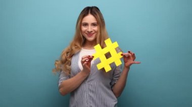Sarı hashtag sembolü taşıyan gülümseyen güzel bir kadın, önemli konuları popüler yapıyor, internete trendler koyuyor, çizgili gömlek giyiyor. Mavi arka planda kapalı stüdyo çekimi.