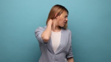 Boynuna dokunan sağlıksız sarışın kadının portresi ağrı ve uyuşukluk, kas gerilimi, osteokondroz, çizgili gömlek giyme endişesi. Mavi arka planda kapalı stüdyo çekimi.