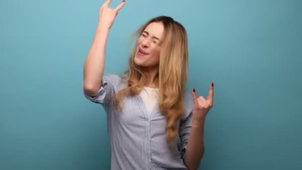 一个喜形于色的女人的画像 她展示了摇滚的手势 尖叫和对重金属的手势 摇滚音乐 穿着条纹衬衫 室内拍摄被蓝色背景隔离 — 图库视频影像