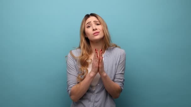 祈りの中で手を取り合っている女性の肖像画 嘆願表現を求めて 助けを請う 許しを求める ストライプのシャツを着ている 青い背景に隔離された屋内スタジオショット — ストック動画