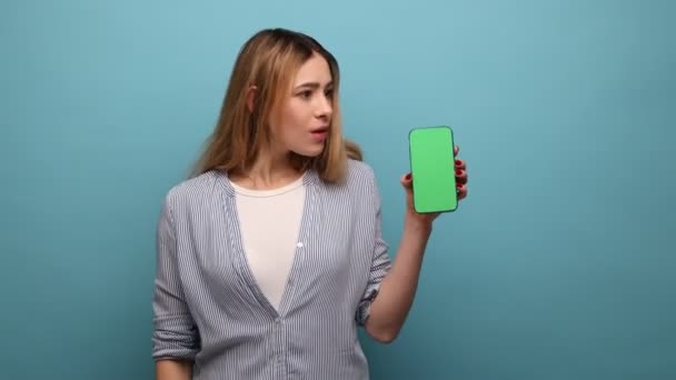 携帯電話を見てみろ 驚くべき女性が指でスマートフォンを指して すごい モバイルデバイスの広告は 縞模様のシャツを着ていると述べた 青い背景に隔離された屋内スタジオショット — ストック動画
