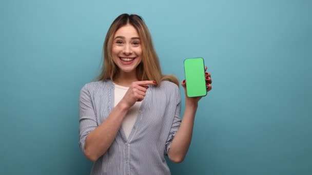 携帯電話を見てみろ スマートフォンを指差し カメラで笑顔 モバイル機器の広告 ストライプのシャツを身に着けている愛らしい幸せな女性 青い背景に隔離された屋内スタジオショット — ストック動画