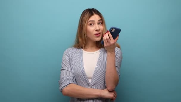 スマートフォンにデジタル音声アプリを使って話している美しい女性の肖像画 メッセージを記録し インターネット上で情報を検索し 縞模様のシャツを着ている 青い背景に隔離された屋内スタジオショット — ストック動画