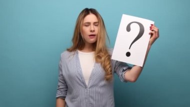 Kafası karışık düşünceli bir kadının kameraya baktığı, elinde soru işareti olan bir kağıt olan portresi görevleri düşünür, çizgili gömlek giyer. Mavi arka planda kapalı stüdyo çekimi.