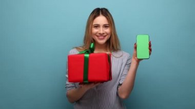 Mobil kullanıcı için ikramiye. Online alışveriş reklamları için boş ekranlı cep telefonu ve hediye kutusu taşıyan mutlu bir kadının portresi, çizgili gömlek giyiyor. Mavi arka planda kapalı stüdyo çekimi