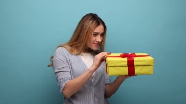 Hediye kutusunu tutan üzgün genç bir kadının portresi, hoşnutsuz bir ifadeyle hediyeyi açıyor, çizgili gömlek giyiyor. Mavi arka planda kapalı stüdyo çekimi.