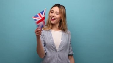 Elinde İngiliz bayrağı sallayan, dişlek tebessümle kameraya bakan, çizgili gömlek giyen iyimser pozitiflerin portresi. Mavi arka planda kapalı stüdyo çekimi.