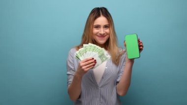 Gülümseyen zengin bir kadının portresi. Elinde akıllı bir telefon, reklam için boş ekranı ve çizgili gömleği olan bir avroluk banknotla. Mavi arka planda kapalı stüdyo çekimi.