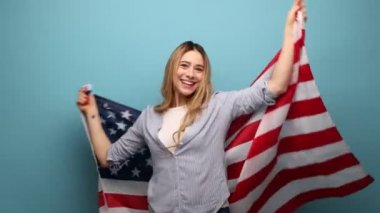 Dalgalı saçlı, neşeli ve çekici bir kadın Amerikan bayrağını omuzlarının üzerinde tutuyor ve mutlu bir şekilde gülümsüyor, ulusal bayramı kutluyor, çizgili gömlek giyiyor. Mavi arka planda kapalı stüdyo çekimi.