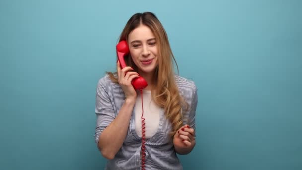 肯定的な幸せな若い女性の肖像画は 幸福を表現し ストライプのシャツを着て 携帯電話で固定固定固定固定固定電話話している 青い背景に隔離された屋内スタジオショット — ストック動画