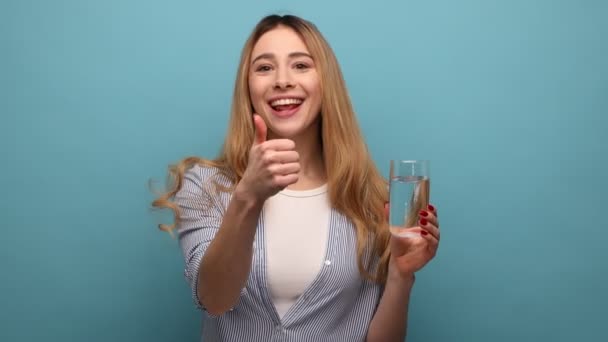 正面女性的画像 手指指向杯中的水 露出大拇指 正面而快乐的表情 穿着条纹衬衫 室内拍摄被蓝色背景隔离 — 图库视频影像