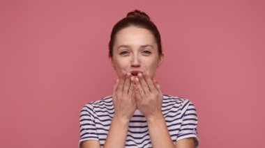 Öpücüğümü yakala. Güzel, romantik bir kadın kameraya bakıyor, avuçlarının üzerinden hava öpücüğü veriyor, aşkı ifade ediyor, çizgili tişört giyiyor. Pembe arka planda kapalı stüdyo çekimleri.