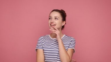 Sevimli, hayalperest, genç bir kadın ayakta duruyor, hayal kurarken yan tarafa bakıyor, dilek tutuyor, olumlu ifade ediyor, çizgili tişört giyiyor. Pembe arka planda kapalı stüdyo çekimleri.