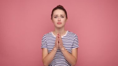 Lütfen, ihtiyacım var. Dua ederken el ele tutuşan ve yalvaran, umut dolu, çizgili tişört giyen genç bir kadın. Pembe arka planda kapalı stüdyo çekimleri.
