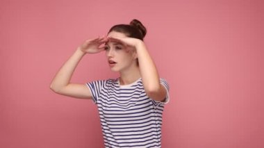 Çekici bir kadın, avuç içini gözlerine bastırıyor. Mesafeli bir bakış açısıyla, ufuk çizgisine bakıyor. İleride, çizgili tişört giyiyor. Pembe arka planda kapalı stüdyo çekimleri.