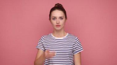 Bun saç stili, ağzı avuç içi ile kaplı, kimseye söylemeyen, çizgili tişört giyen güzel, genç bir kadın. Pembe arka planda kapalı stüdyo çekimleri.