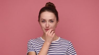 Kurnaz bir kadının kameraya bakması ve ağızını eliyle kapatması, dudaklarını fermuarlaması, yani kimseye söylemeyeceğim, çizgili tişört giyeceğim. Pembe arka planda kapalı stüdyo çekimleri.