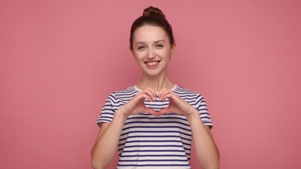 幸せな美しい笑顔の女性の愛のジェスチャーで立って 心臓の形のジェスチャーでカメラを見て 縞模様のTシャツを着て ピンクの背景に隔離された屋内スタジオショット — ストック動画