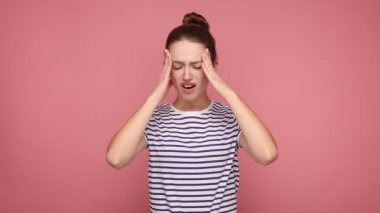 Kıllı topuzlu bir kadın, elleri başı ağrıyan bir kadın, şiddetli ağrıdan çatık kaşları çatıyor, dayanılmaz migren ağrısı, stres gerilimi, çizgili tişört giyiyor. Pembe arka planda kapalı stüdyo çekimleri.