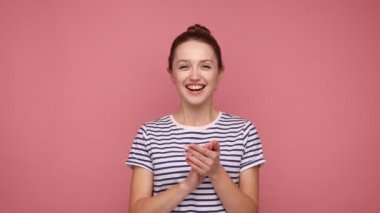 Bun saç stili alkışlayan, alkışlayan, zaferle el çırpan çizgili tişört giyen neşeli, genç bir kadın. Pembe arka planda kapalı stüdyo çekimleri.