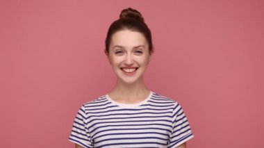 Mutlu, iyimser, çekici, saçları topuzlu bir kadın evet diyor, anlaşmayı ifade ediyor, kameraya olumlu bir ifadeyle bakıyor, çizgili tişört giyiyor. Pembe arka planda kapalı stüdyo çekimleri.
