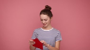 Gülümseyen, saçları topuzlu güzel bir kadın hassas bir mektupla, okumayla, romantik bir mesajla, çizgili tişörtle dokunuluyor. Pembe arka planda kapalı stüdyo çekimleri.