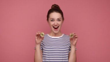 Bun saç stiline sahip son derece mutlu bir kadın altın bitcoin ile kaplı bir göz, engelleme zinciri konsepti, heyecan ifade eden, çizgili tişört giyen. Pembe arka planda kapalı stüdyo çekimleri.