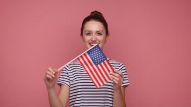 Amerikan bayrağını tutan, Amerikan bayrağını taşıyan çekici bir kadın. Ulusal bayramı kutlayan, kameraya bakan, çizgili tişört giyen. Pembe arka planda kapalı stüdyo çekimleri.