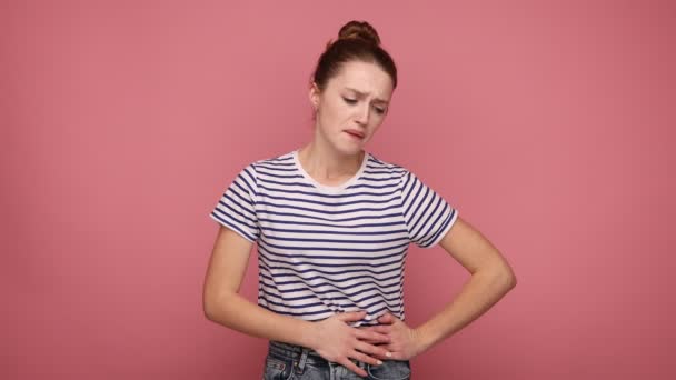 動揺の女性は ひどい急性の痛み 虫垂炎の炎症の症状 流産のリスクに苦しんで 縞模様のTシャツを着て彼女の腹を保持 ピンクの背景に隔離された屋内スタジオショット — ストック動画