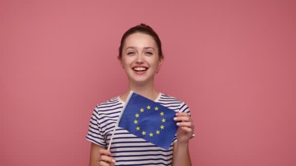 在欧盟的公民身份 快乐的年轻女性 留着束发 展示欧盟国旗 对着相机微笑 穿着条纹T恤 在粉色背景下被隔离的室内拍摄 — 图库视频影像