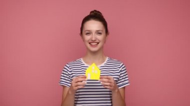 Bun saç stili, neşeli bir yüzü olan, kendi evini düşleyen, çizgili tişört giyen çekici bir kadın. Pembe arka planda kapalı stüdyo çekimleri.