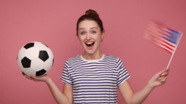 ABD ve futbol topunun bayrağını elinde tutan son derece mutlu bir kadın şampiyonada favori takımı destekliyor, neşeli, çizgili tişört giyiyor. Pembe arka planda kapalı stüdyo çekimleri.