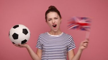 Birleşik Krallık ve futbol topunun bayrağını elinde tutan heyecanlı bir kadın şampiyonada favori takımı destekliyor, keyifli, çizgili tişört giyiyor. Pembe arka planda kapalı stüdyo çekimleri.