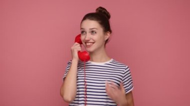 Dost canlısı bir kadın. Sabit kırmızı bir cep telefonu tutuyor. Hoş sohbetler ediyor, gülümsüyor, çizgili tişört giyiyor. Pembe arka planda kapalı stüdyo çekimleri.