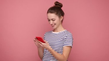 Akıllı telefondaki sosyal ağ paylaşımlarına güler yüzlü bir kadın, cep telefonundan memnun, çizgili tişört giyen bir kullanıcı. Pembe arka planda kapalı stüdyo çekimleri.