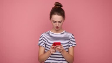 Şok olmuş bir kadın, sosyal ağda saç teli okuyup cep telefonu kullanıyor, sohbet ediyor, şaşırmış görünüyor, çizgili tişört giyiyor. Pembe arka planda kapalı stüdyo çekimleri.