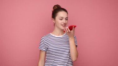 Akıllı telefonla konuşan pozitif bir kadın, sanal asistan, ai akıllı teknoloji, ses uygulaması, sesli mesaj, çizgili tişört giyiyor. Pembe arka planda kapalı stüdyo çekimleri.