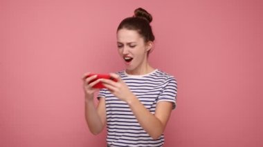 Mutlu heyecanlı kadın cep telefonu kullanıyor ve gülümsüyor, cep telefonuyla video oyunu oynuyor, heyecanını dile getiriyor, çizgili tişört giyiyor. Pembe arka planda kapalı stüdyo çekimleri.