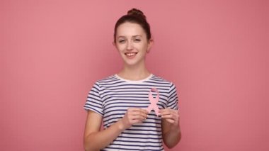 Hey sen, sağlığına dikkat et. Güzel bir kadın pembe kurdele tutuyor, göğüs kanseri farkındalığının sembolü, kamerayı işaret ediyor, çizgili tişört giyiyor. Pembe arka planda kapalı stüdyo çekimleri.