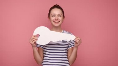 Tüylü olumlu bir kadın büyük kağıt anahtarı işaret ediyor ve kameraya dişlek gülüşüyle bakıyor, satın alıyor, baş parmağını gösteriyor, çizgili tişört giyiyor. Pembe arka planda kapalı stüdyo çekimleri.