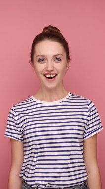 Şaşkınlık ve şaşkınlık ifade eden, büyük gözlü kameraya bakan, çizgili tişört giyen genç bir kadının dikey görüntüsü. Pembe arka planda dikey stüdyo çekimi.