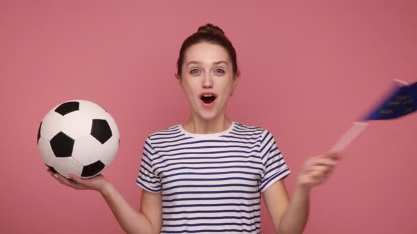 ヨーロッパの国旗とサッカーボールを手にした現役の幸せな女性は 選手権でお気に入りのチームをサポートし ストライプのTシャツを着ています ピンクの背景に隔離された屋内スタジオショット — ストック動画