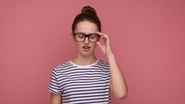 迷人疲惫的女人拿着发髻摘下眼镜 感到眼睛疼痛 因长时间工作而疲倦 穿着条纹T恤 在粉色背景下被隔离的室内拍摄 — 图库视频影像