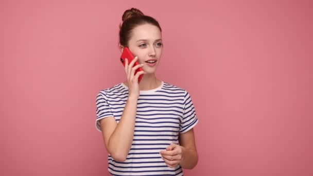 陽気な女性の携帯電話で友人と話し 離れて見て 楽しく笑顔 楽しい会話をし 縞模様のTシャツを着ている ピンクの背景に隔離された屋内スタジオショット — ストック動画