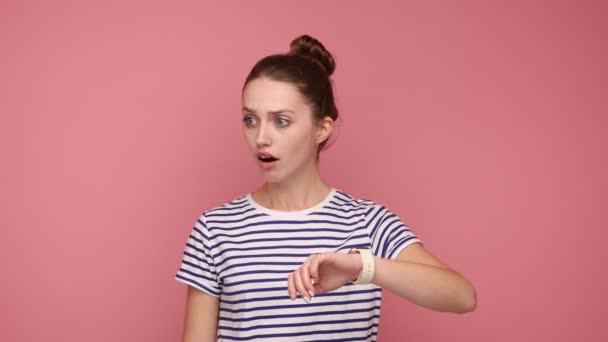 髪パン指の爪をかむと彼女の手首の時計を見て 期限については 縞模様のTシャツを着て心配して神経の女性 ピンクの背景に隔離された屋内スタジオショット — ストック動画